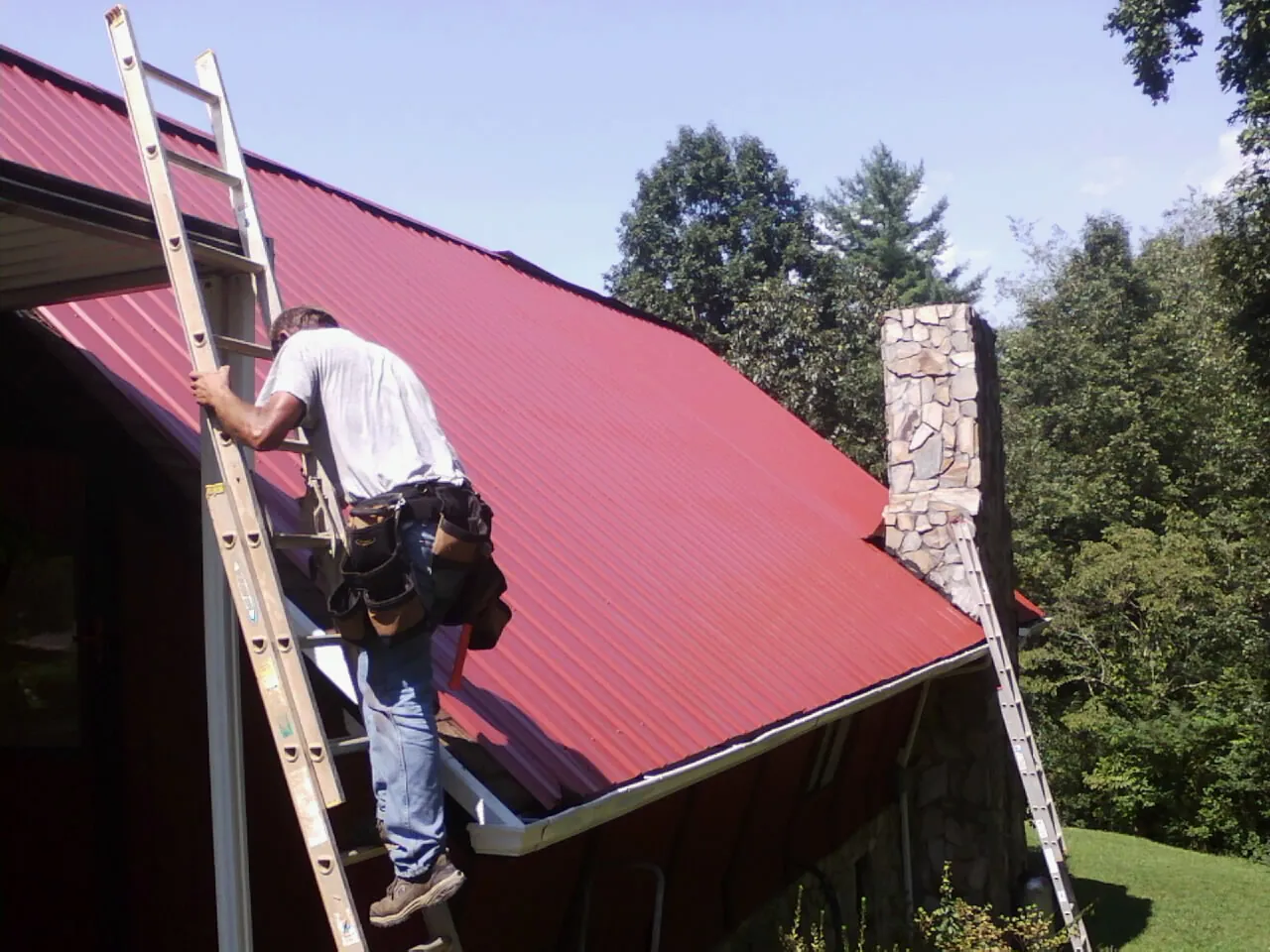 Roofer on a ladder beside a red metal roof and stone chimney — Soffit & Fascia Repair by Roofing Brothers in Mechanicsburg